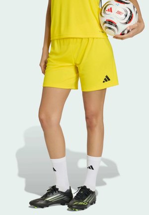 Osoba ubrana w żółte sportowe spodenki i koszulkę, białe skarpetki Adidas, czarne korki, trzymająca biało-czerwoną piłkę nożną Adidas.