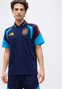 Jaunas vyras dėvi tamsiai mėlyną sportinį marškinėlį ir šortus su raudonais, geltonais ir šviesiai mėlynais akcentais, kuriuose yra Ispanijos nacionalinės futbolo komandos emblema.