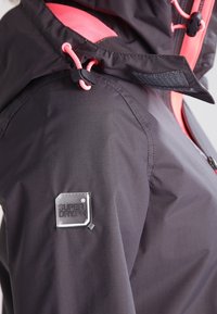 Detailní záběr ramene černé a růžové nepromokavé bundy s nastavitelným šňůrkou na kapuci a nášivkou s logem Superdry.