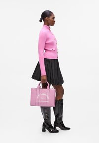Roze geribbelde trui met gouden knopen, zwarte flared rok en glanzende hoge zwarte laarzen. Draagt een roze handtas met witte branding.