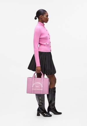 Pink ribbed sweater with gold buttons, black flared skirt, and glossy knee-high black boots. Carrying a pink handbag with white branding.