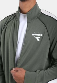 Giacca verde oliva con zip, realizzata in tessuto morbido, caratterizzata da un collo alto, strisce laterali bianche e un logo bianco sul petto.