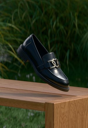 Schwarzer Lederloafer mit goldener Kettendetail, der über einer Holzbank im Freien schwebt, mit grünem Gras und Pflanzen im Hintergrund.
