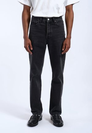 Man wearing straight-leg black jeans, white short-sleeve shirt, and black loafers standing against a plain white background.