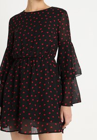 Vestido negro con estampado de corazones rojos, mangas acampanadas transparentes y cintura ceñida, llevado por una persona mostrada desde el cuello hasta la mitad del muslo.