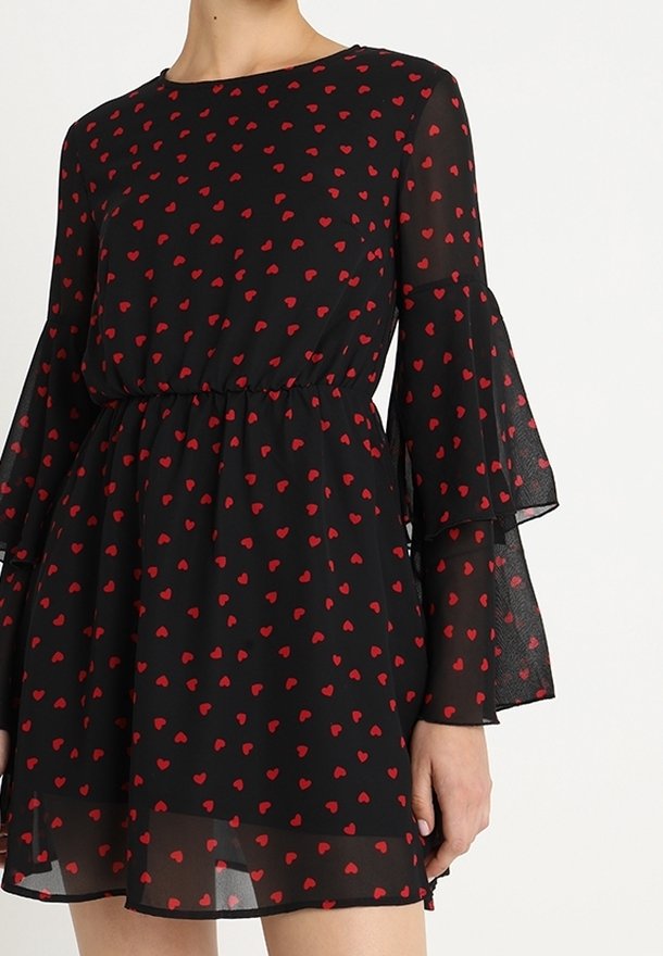 Vestido negro con estampado de corazones rojos, mangas acampanadas transparentes y cintura ceñida, llevado por una persona mostrada desde el cuello hasta la mitad del muslo.