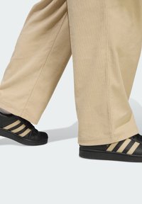 Black sneakers with three beige stripes, textured leather, and rubber soles, paired with wide, ribbed beige corduroy pants.