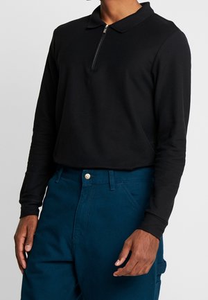 Mann trägt ein schwarzes langärmliges Polohemd mit Reißverschluss und blaue Hosen, steht vor einem schlichten hellgrauen Hintergrund.