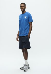 Blue cotton t-shirt with a white logo on the chest, paired with navy shorts and gray athletic shoes. Model is standing against a plain background.