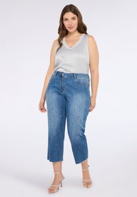Top senza maniche grigio chiaro, jeans cropped in denim blu con decorazioni in perline colorate e tacchi argentati con cinturini. Dettaglio con orlo sfrangiato sui jeans.