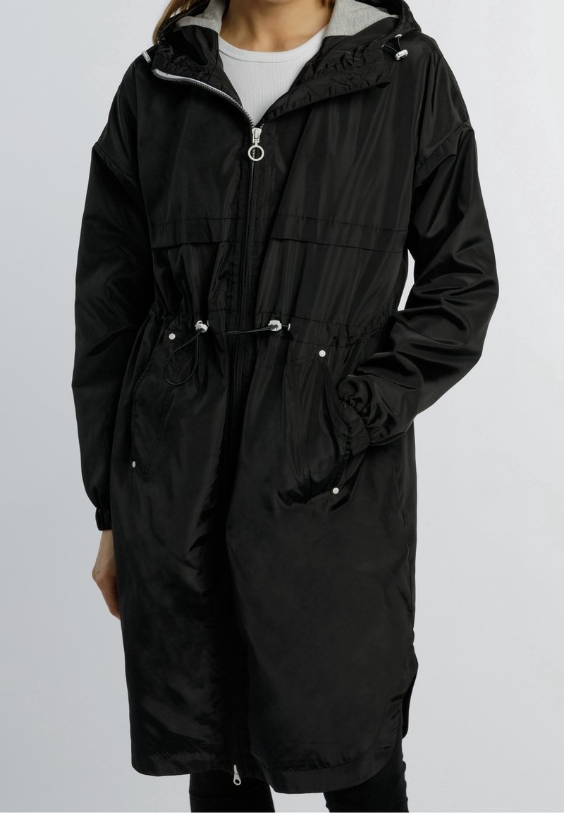 Schwarzer langer Mantel aus leichtem Stoff, mit Reißverschluss vorne, elastischem Kordelzug in der Taille und zwei Seitentaschen. Die Kapuze ist grau gefüttert.