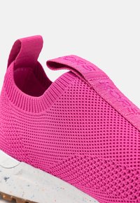 Gros plan d'une basket slip-on tricotée rose vif avec languettes et semelle blanche tachetée.