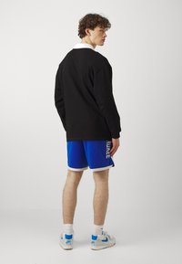 Camisa negra de manga larga con cuello blanco, pantalones cortos deportivos azules con un ribete plateado y logotipo, y zapatillas blancas con acentos azules.