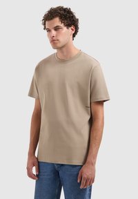Beige katoenen t-shirt met korte mouwen, ronde hals, en een klein zwart "PURE" label aan de linkerkant nabij de zoom. Draagt met blauwe jeans.