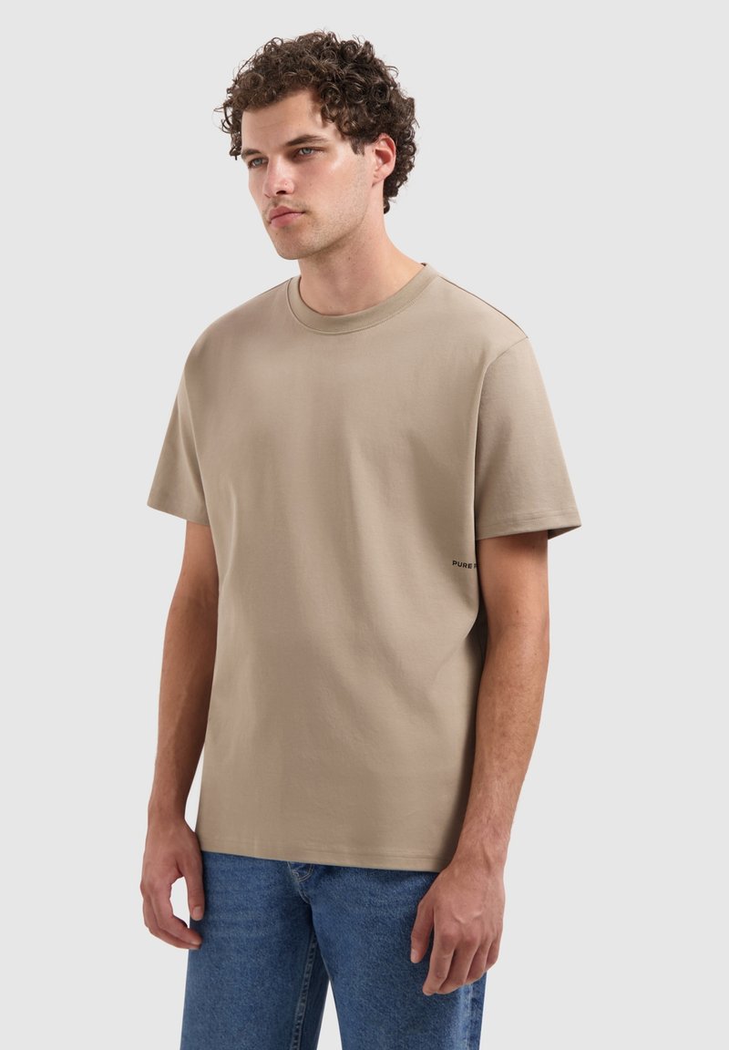Beige katoenen t-shirt met korte mouwen, ronde hals, en een klein zwart "PURE" label aan de linkerkant nabij de zoom. Draagt met blauwe jeans.