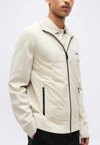 Άνδρας που φοράει μπεζ ζακέτα με φερμουάρ και τσέπες σε μαύρο χρώμα, με το λογότυπο "BOSS" στο στήθος και μπεζ πόλο μπλούζα από κάτω.