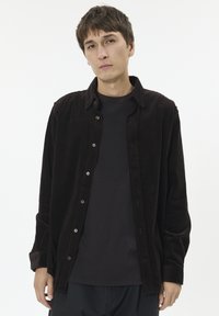 Brun kordfløjlsskjorte med knapfront, med en spredt krave og lange ærmer, lagd ovenpå en mørkstribet t-shirt.