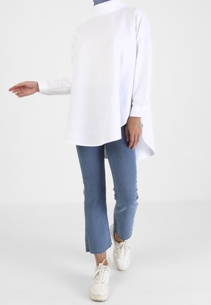 Personne portant un chemisier blanc asymétrique à manches longues, un jean bleu court et des baskets blanches, avec un bras légèrement tendu.