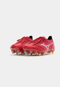 Červené futbalové kopačky s textúrovaným zvrškom, s čiernymi akcentmi a bielym logom. Priehľadné cleats s oranžovými špičkami na podrážke.