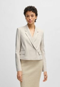 Lichtbeige cropped blazer met opstaande revers, twee vo zakken en een textuurstof, gedragen over een geribbelde beige rok.