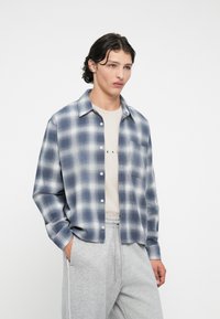 Flanelasta srajca z modro-belim karo vzorcem, z gumbi spredaj, široko ovratnico in enim prsnim žepom, nošena čez bež srajco.