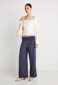 Balts cieši pieguļošs garroku tops kopā ar tumši ziliem platajiem biksēm ar savelkamu jostu; modelim rokā kremkrāsas soma un nude toņa dzīvokļi.