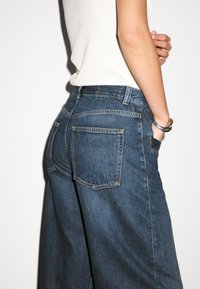 Jeans in denim blu scuro con vita alta, taglio a gamba larga e due tasche posteriori. Presentano cuciture visibili e un classico design a cinque tasche.