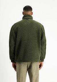 Oliwkowa polarowa kurtka z wysokim kołnierzem, teksturowana powierzchnia i elastyczne mankiety. Gładkie akcenty z tkaniny na rękawach i bokach.