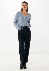 Kobieta stojąca z rękami w kieszeniach, ubrana w jasnoniebieski dziergany kardigan zapinany na guziki, czarne spodnie o wysokim stanie i czarne botki za kostkę na białym tle.