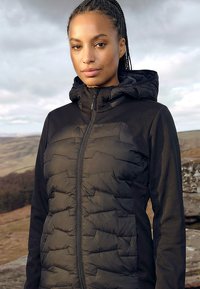 Nő, akinek befont haja van, fekete steppelt kapucnis dzsekit visel, szabadtéren áll, háttérben dombok és felhős égbolt látható.