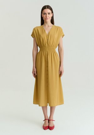 Femme debout, face à l’avant, portant une robe midi boutonnée jaune moutarde et des chaussures plates rouges à bride cheville sur un fond uni.