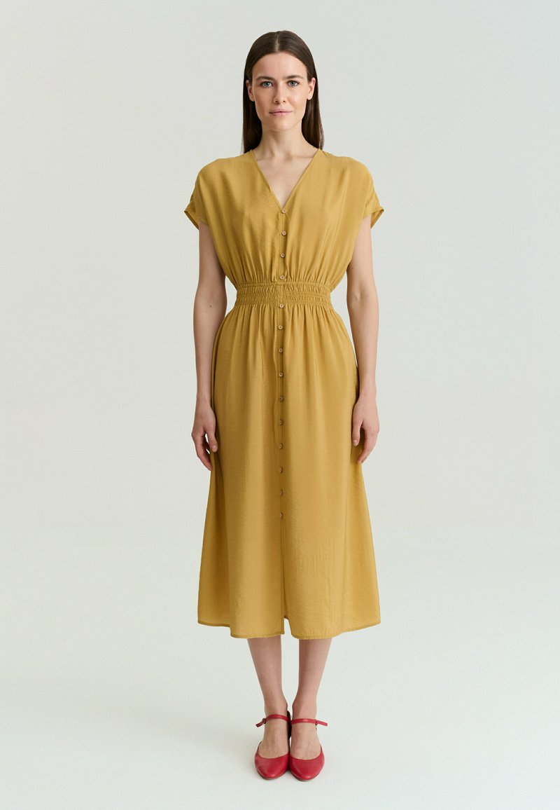 Femme debout, face à l’avant, portant une robe midi boutonnée jaune moutarde et des chaussures plates rouges à bride cheville sur un fond uni.