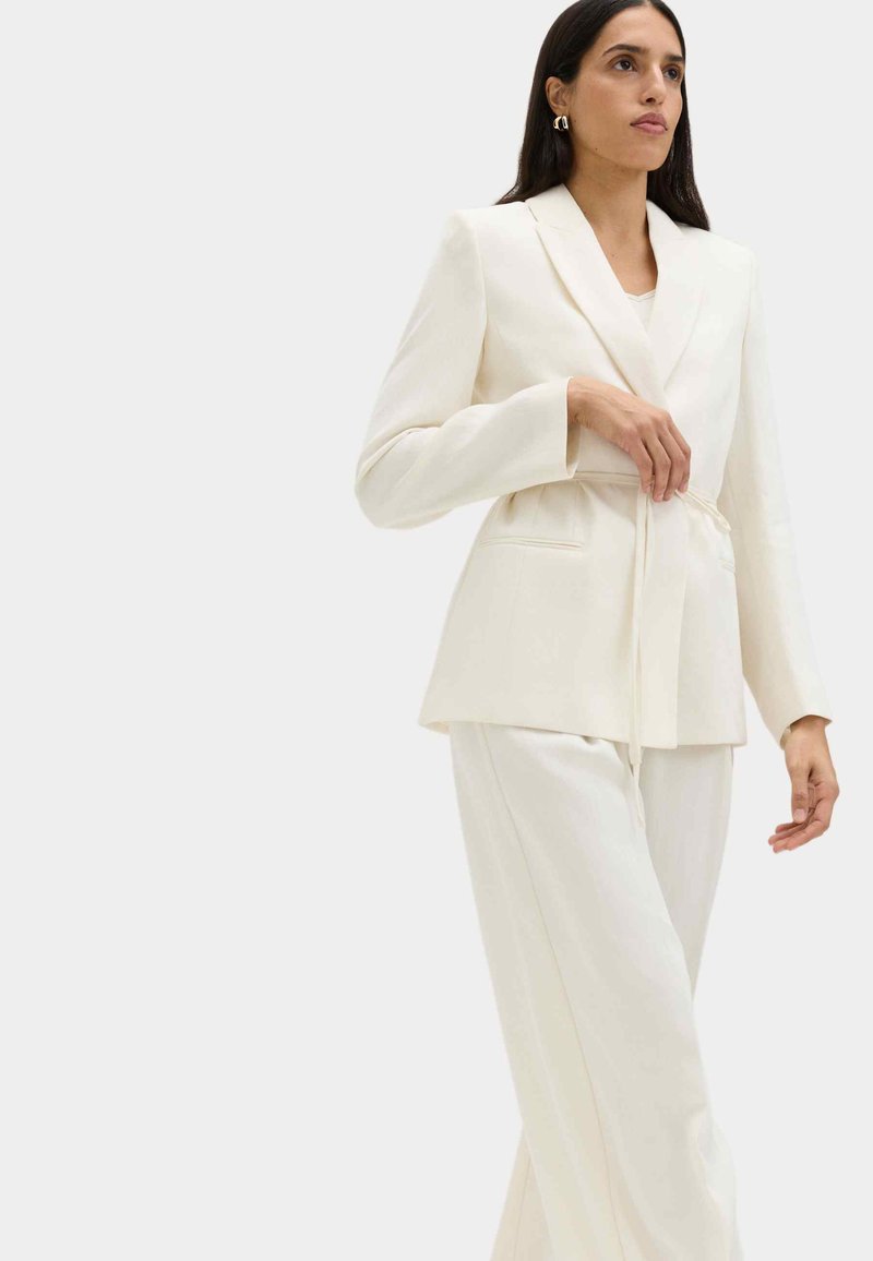Woman with long dark hair adjusting the tie of an off-white tailored blazer, paired with matching wide-leg trousers, on light background.