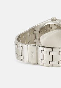Srebrny zegarek metalowy o polerowanej powierzchni, z bransoletą w linki i kwadratową kopertą. Zapięcie ma wygrawerowane logo "GUESS".