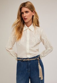 Chemise blanche à manches brodées, col classique, associée à un jeans en denim bleu taille haute et une ceinture en corde tressée avec des franges.