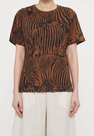 Persona con camiseta de manga corta de estampado de cebra en marrón y negro y pantalones sueltos de color beige claro, de pie contra un fondo liso.