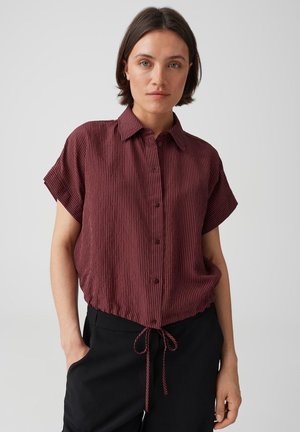 Donna con capelli corti castani che indossa una camicetta a righe bordeaux a maniche corte con bottoni e pantaloni neri con le mani nelle tasche.