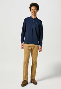 Marineblauw poloshirt met lange mouwen en drie-knopen-voorpand, gedragen met tan corduroy broek en bruine schoenen; soepel stof, casual pasvorm.