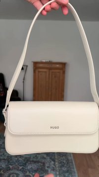 Χέρι κρατάει μια λευκή τσάντα ώμου Hugo με καμπυλωτό ιμάντα μπροστά σε ένα δωμάτιο με ξύλινη ντουλάπα και χαλί ορατά.
