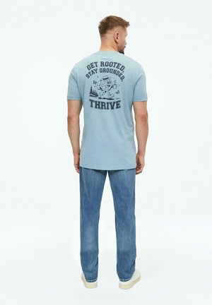 Mann trägt hellblaues T-Shirt mit Wander-Cartoon und Text "Get Rooted Stay Grounded Thrive" auf dem Rücken, blaue Jeans und weiße Schuhe.