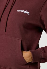 Bordeaux hoodie met een voorzak, capuchon met trekkoord en een wit "wrangler" logo. Gemaakt van zachte, gestructureerde stof met ribgebreide manchetten.
