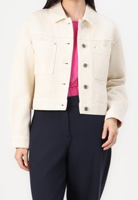 Chaqueta de denim crema con cuello, bolsillos frontales y botones plateados, combinada con una blusa rosa y pantalones oscuros.
