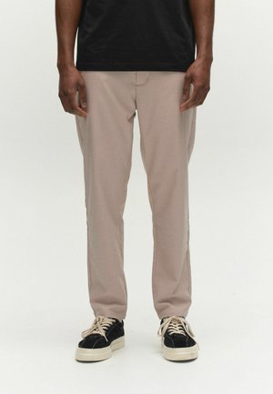 Mann trägt beige Hose, schwarzes Hemd und schwarze Sneakers mit weißen Sohlen und Schnürsenkeln, steht vor einem weißen Hintergrund.