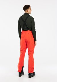 Pantalons de ski orange vif avec bretelles noires réglables, aérations zippées sur les côtés et accents noirs au niveau des chevilles ; le matériau semble durable.