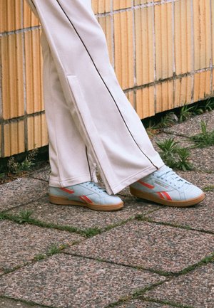 Personne portant un pantalon beige clair et des baskets Reebok bleu clair avec des rayures orange, debout sur un pavé en pierre près d'un mur de briques.