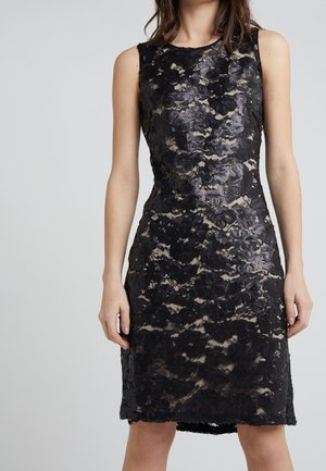 Robe noire sans manches en dentelle avec motif floral sur fond nude, taille cintrée, longueur au genou, portée par une femme debout aux bras détendus.