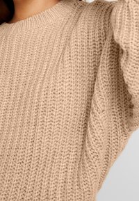 Pull en tricot beige présentant un motif texturé et des poignets côtelés, avec un col rond et un matériau en fil doux et épais.