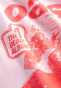 Bela majica s rdečim grafičnim dizajnom. Besedilo vključuje »#1 THE DEBUT ALBUM« ter ilustracijo ljudi in pohištva v prostoru.