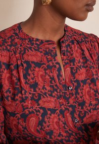 Blusa floreale rossa e blu navy con collo abbottonato, tessuto strutturato, morbide pieghe e colletto classico. Presenta intricati motivi paisley.