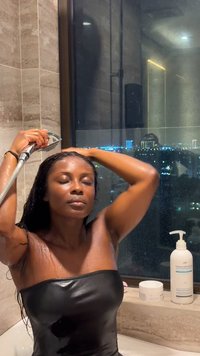 Svart stroppeløs topp, vått hår, dusjhodet holdt opp; bakgrunnen viser en bysilhuett om natten. I nærheten står flasker med sjampo og balsam.